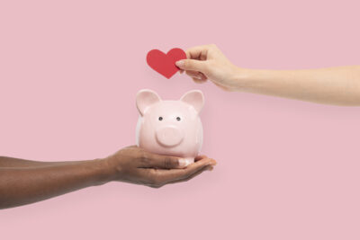 Eine Frau hält ein rosa Sparschwein während eine andere Frau ein rotes Herz aus Papier darüber hält